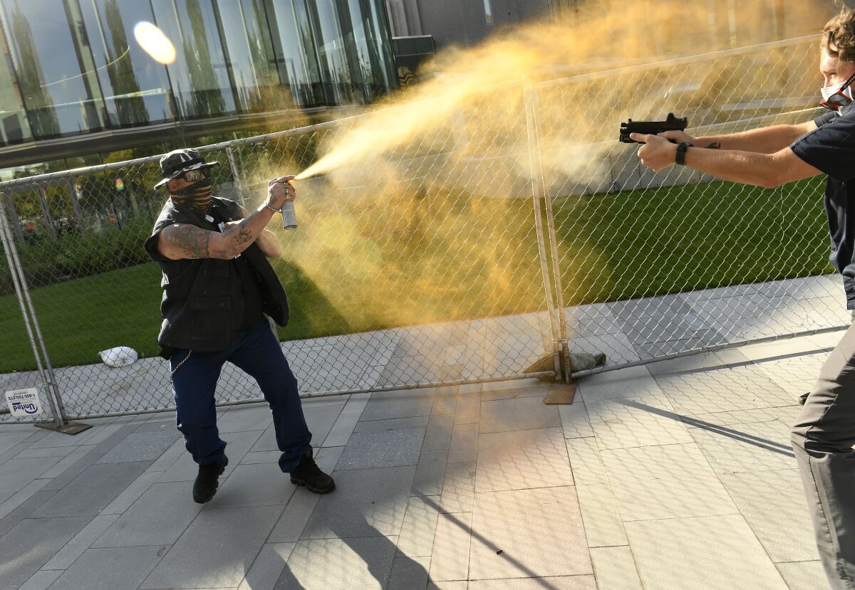 A man in a U.S. flag mask sprays Mace at a person pointing a handgun at him