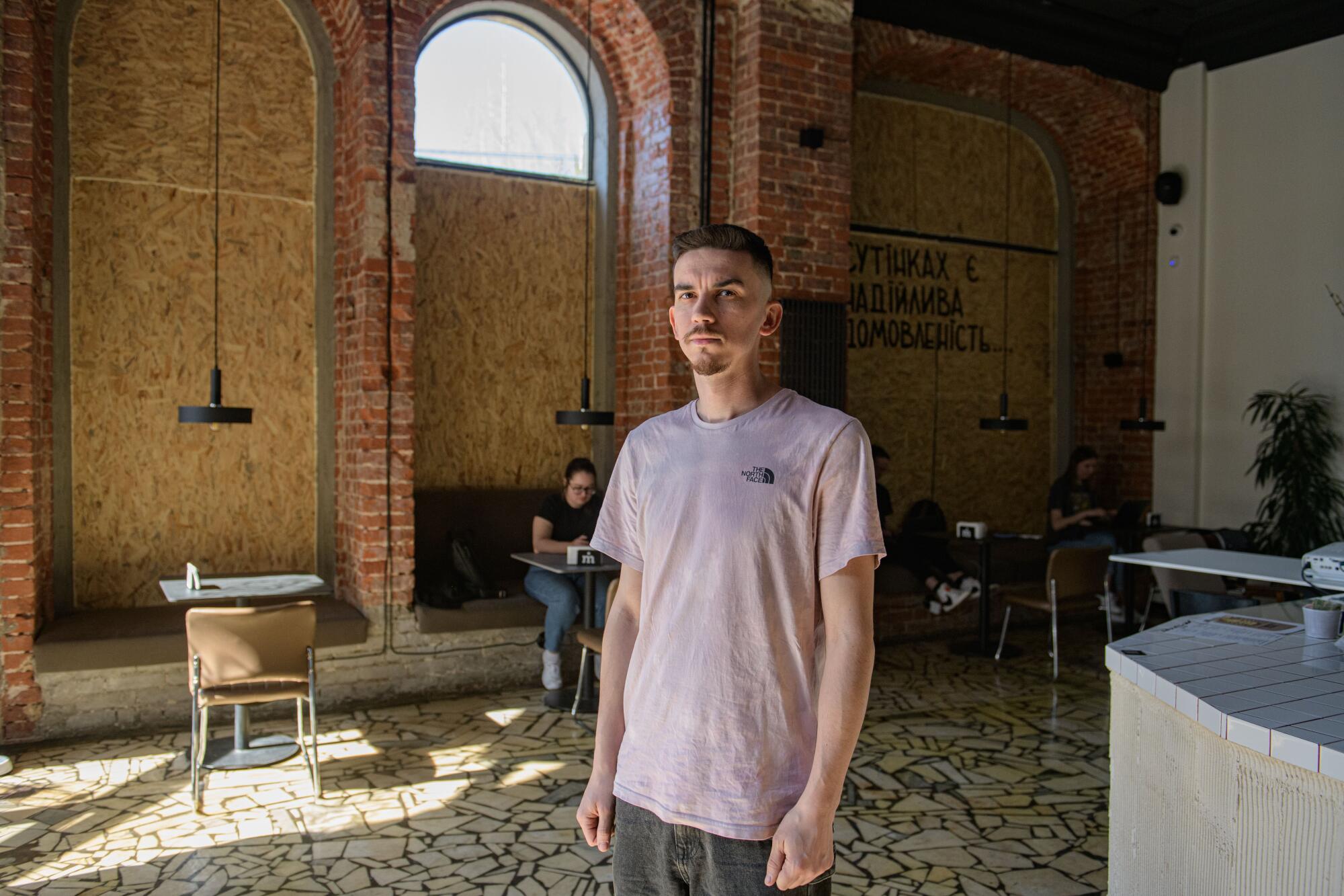 A bearded man in beige T-shirt stands in a cafe with brick walls and boarded-up windows 