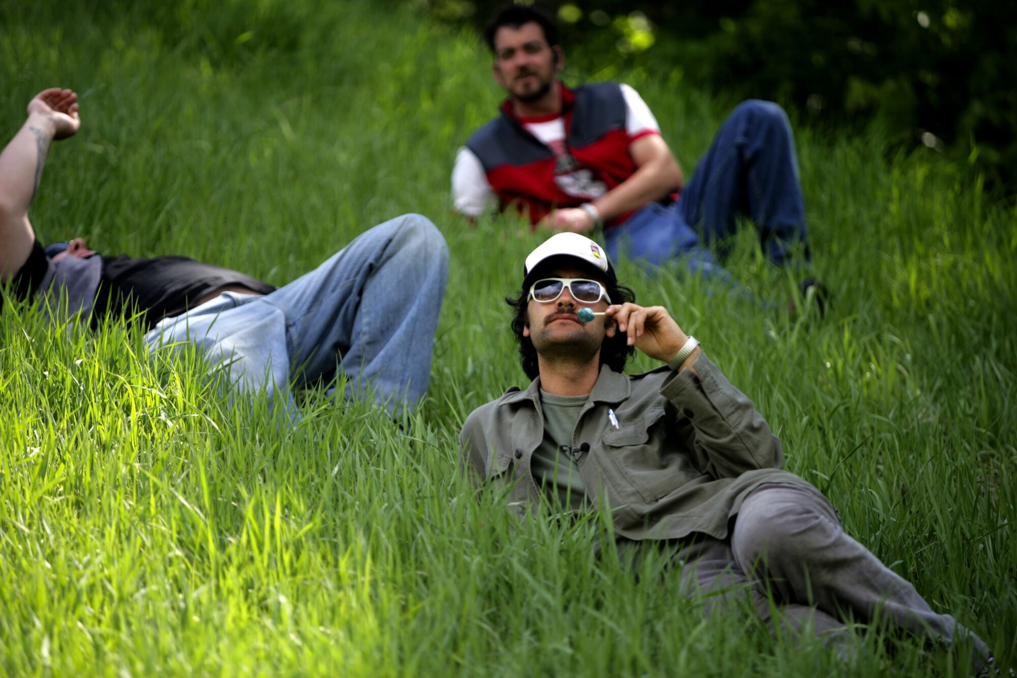 A man with a lollypop lies in the grass with two other men in the background