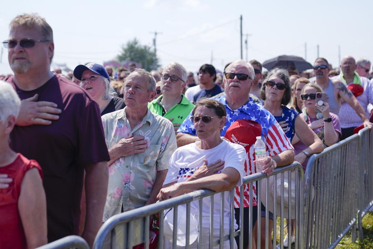 Rally attendees stand near a metal barrier with their right hands on their chests during the Pledge of Allegiance.