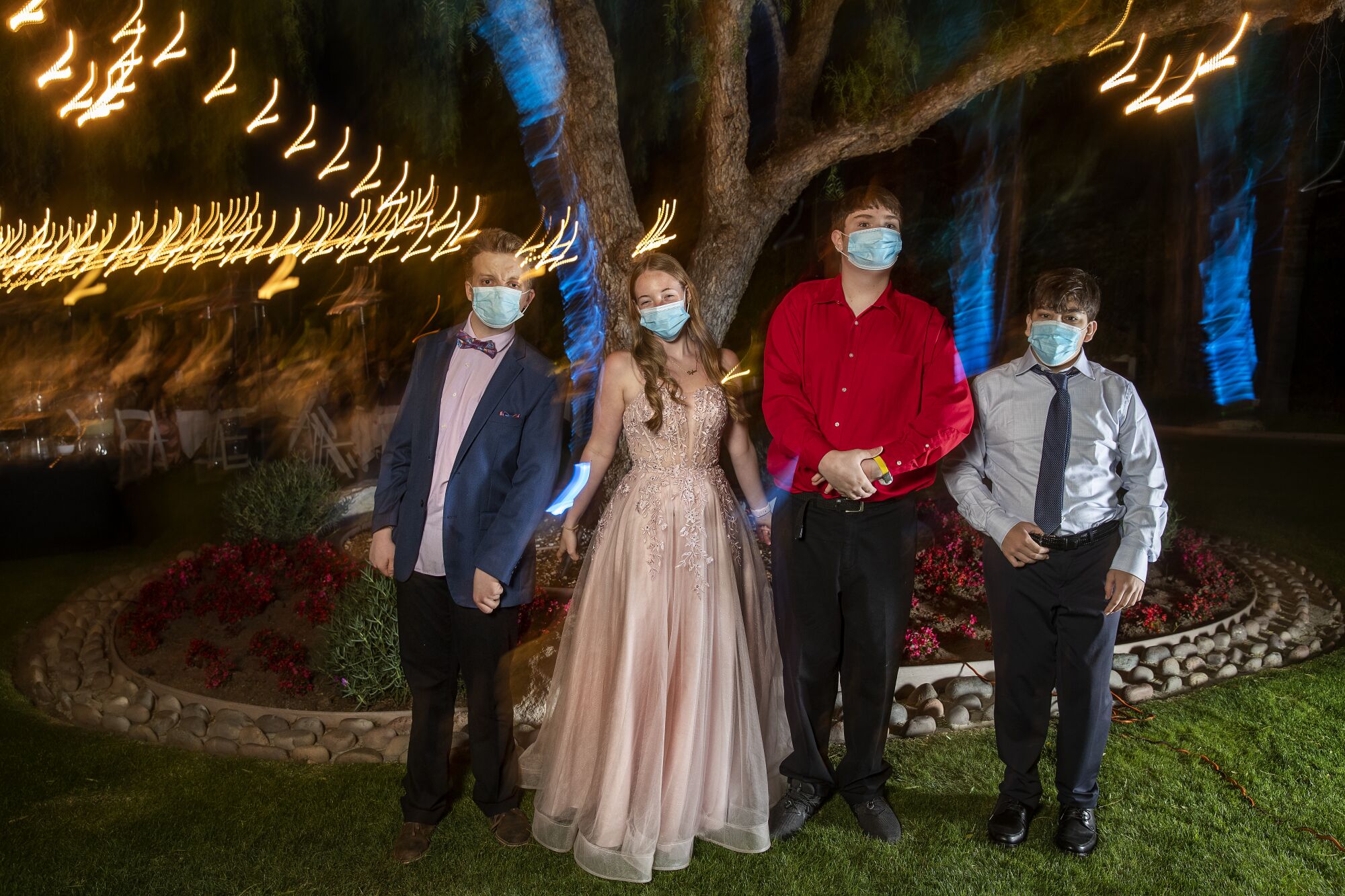 Students in formal wear and masks in front of a tree with lights.