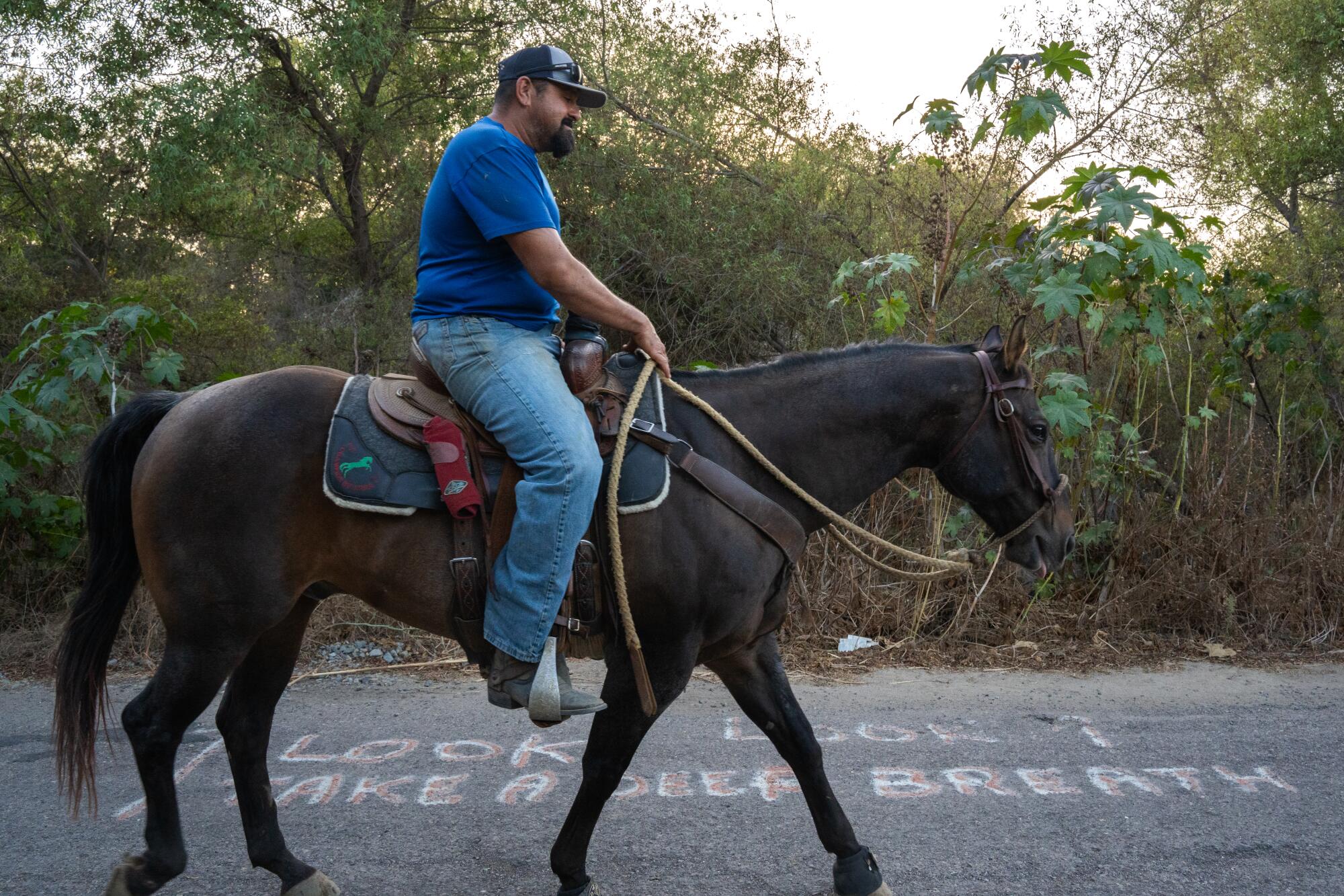 A rider walks his horse on pavement crossing over the Tijuana River. The asphalt reads, "Look, take a deep breath." 