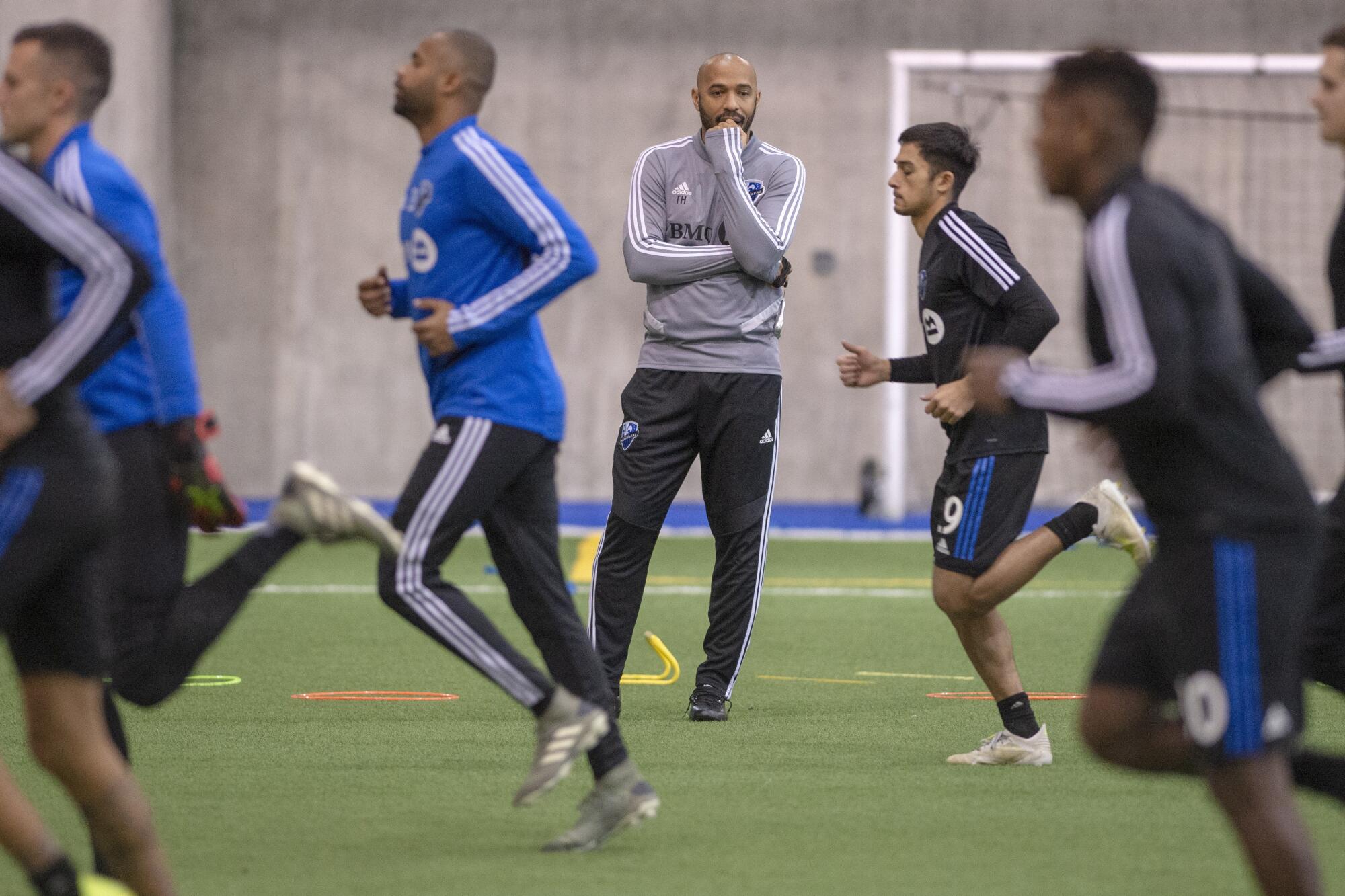 Thierry Henry standing in the middle of a soccer field with players running past him at a practice