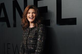 Linda Evangelista smiles while wearing a black patterned coat and skirt against a black backdrop.