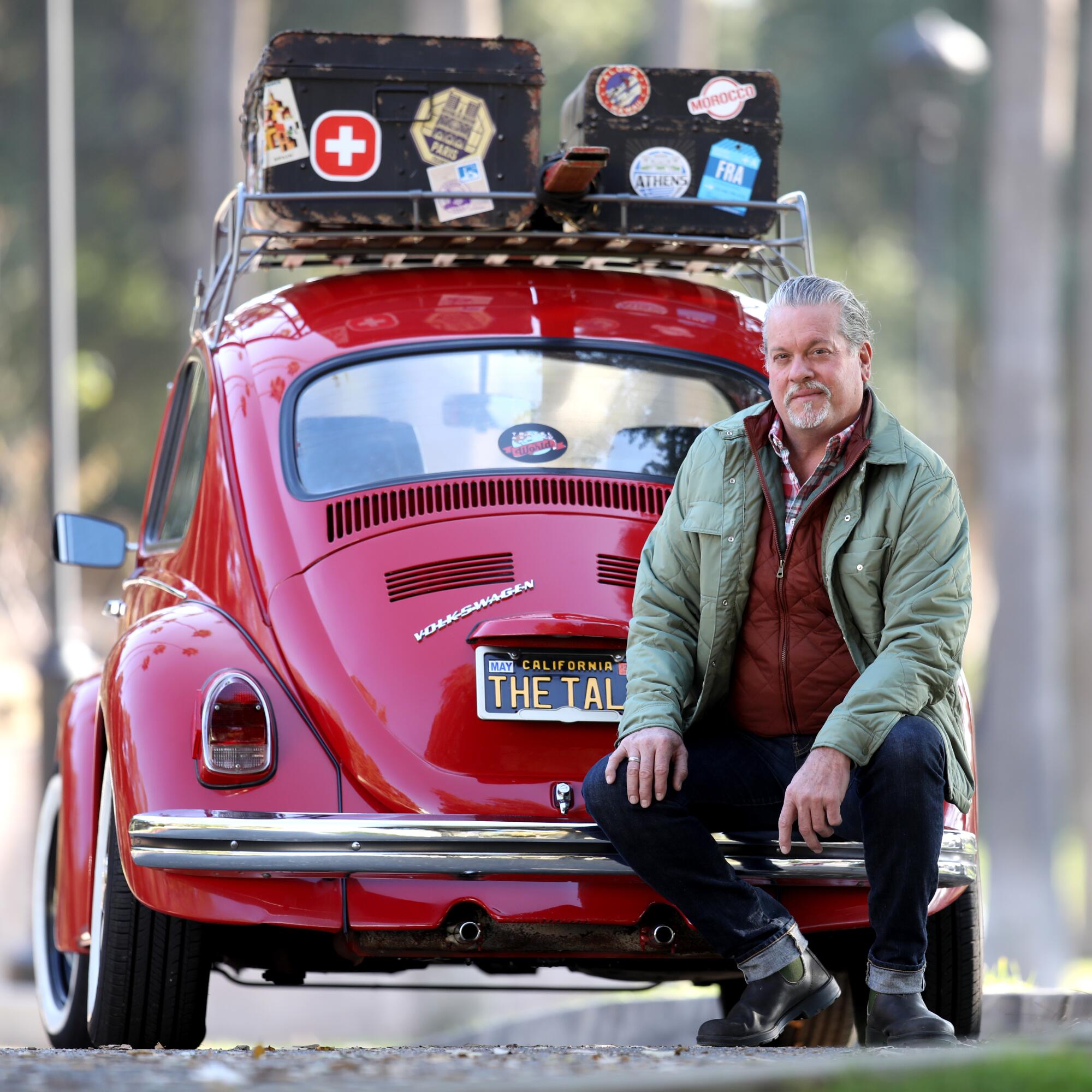 An older man sits on the back bumper of a red Volkswagen Beetle car with sticker-covered boxes on top.