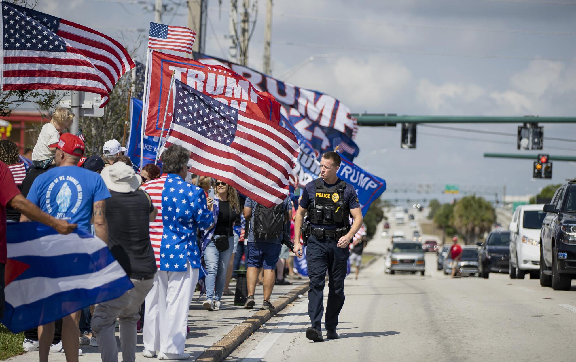 A police officers walks along a row of people standing by a road with U.S. flags and Trump flags.