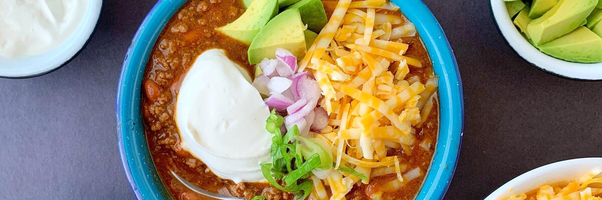 A bowl of beef and bean chili with garnishes of grated cheese, avocado, sour cream, chopped red onion and sliced scallions.