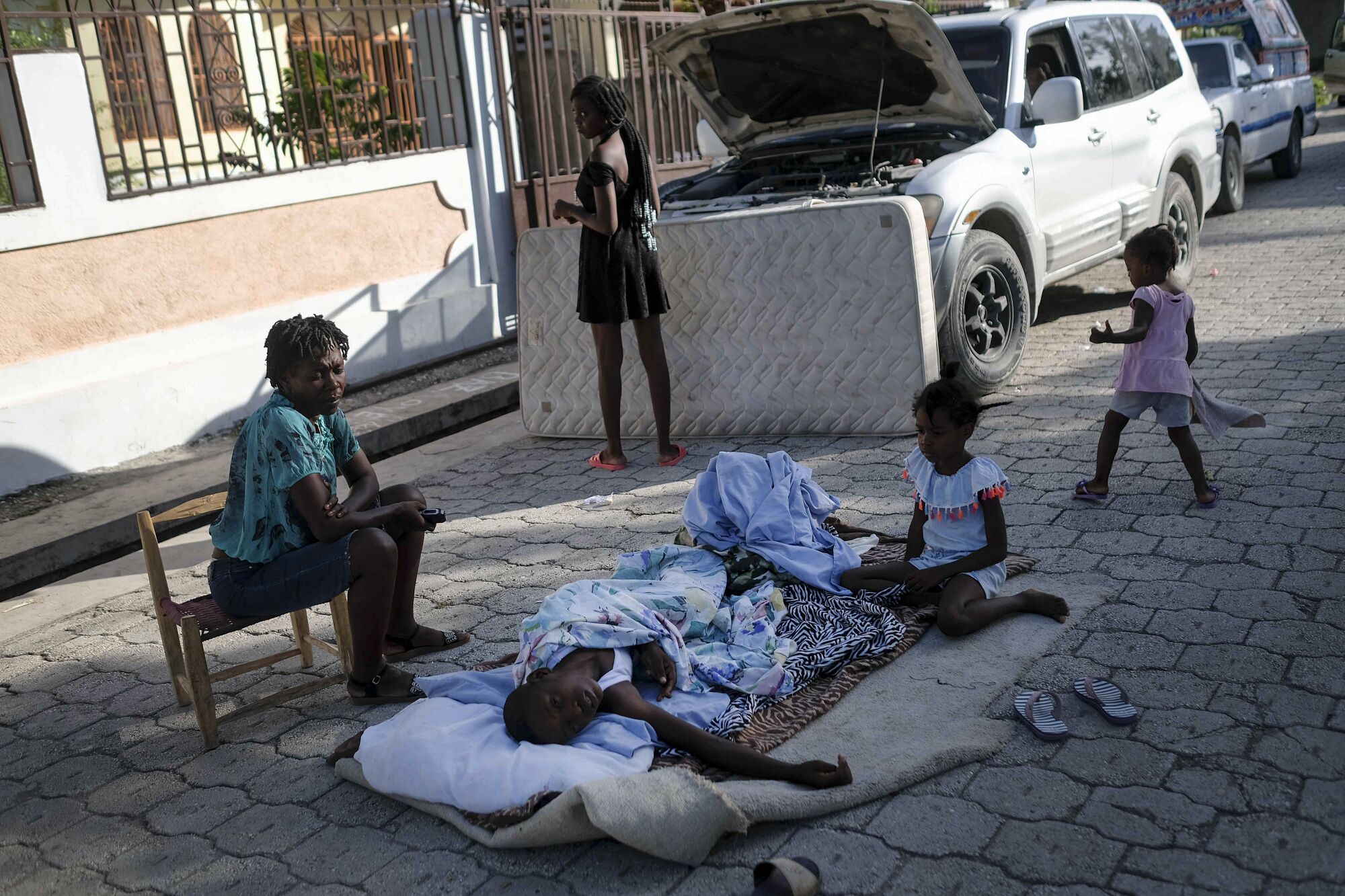 A person lies on bedding in a street while other people stand or sit nearby