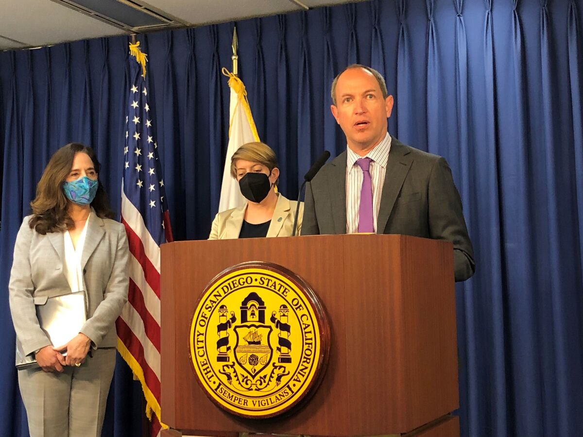 A man in a suit speaks at a podium as two women in masks stand behind him.