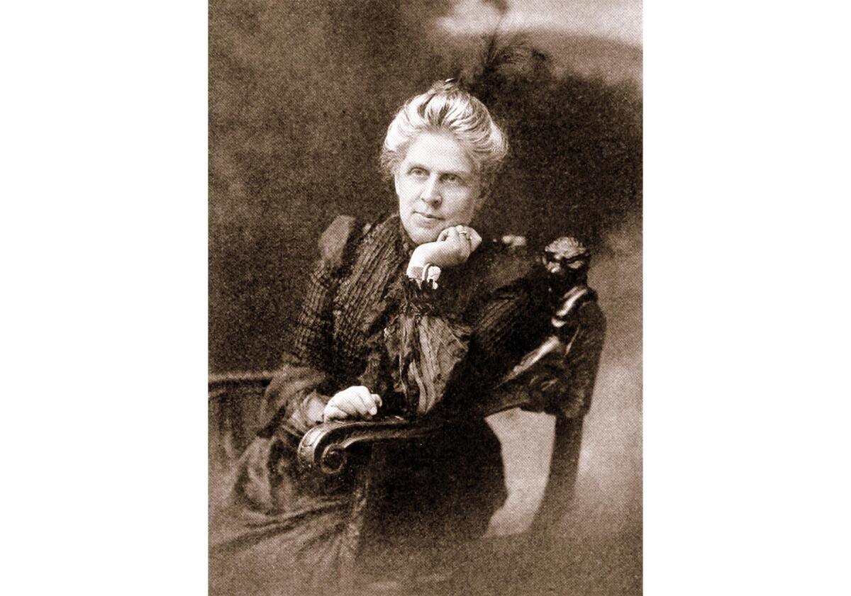 A woman with upswept hair and wearing a long, dark, high-necked dress sits in a chair for a portrait.
