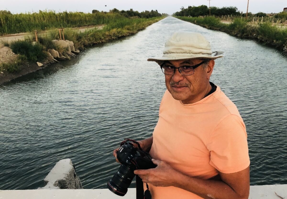 A man wearing a bucket hat and carrying a digital camera stands for a photo against a backdrop of a large river.