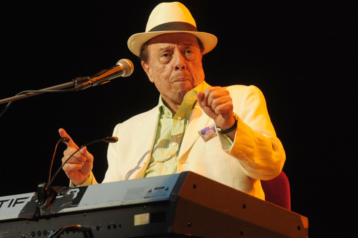 Sergio Mendes in a white suit and a hat holding up his hands while sitting at a keyboard