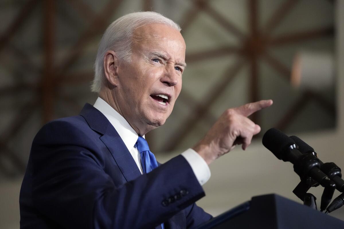 Biden, wearing a dark blue suit, a white shirt and a blue tie, stands at a lectern, pointing with his right hand as he speaks