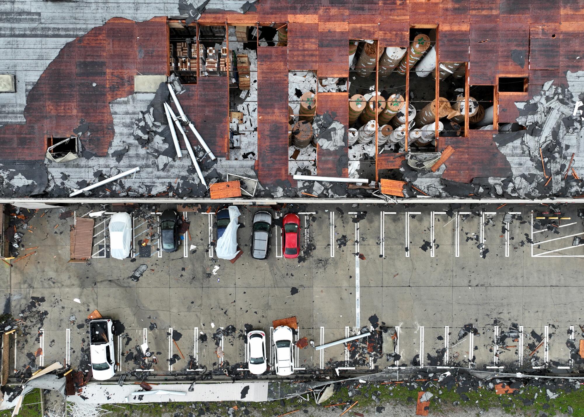 An aerial view of a building with part of its roof missing, exposing the interior, and a nearby parking lot with debris.