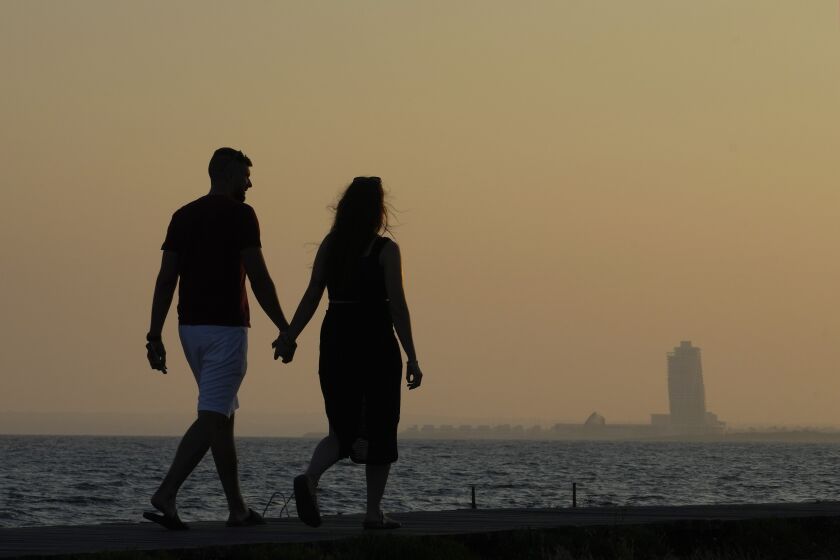 A couple walk by the sea as in the background is seen an apartments tower of a new construction of "Ayia Napa Marina", during sunset in the resort of Ayia Napa in the eastern Mediterranean island of Cyprus, on Tuesday, June 6, 2023. When the U.S. and U.K. in April included a handful of Cypriot nationals and Cyprus-registered companies as part of another global crackdown on 'enablers' helping Russian oligarchs skirt sanctions, the perception that the island nation somehow remains Moscow's financial lackey again loomed large. (AP Photo/Petros Karadjias)