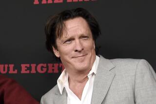 Michael Madsen leans to his right and smiles while waring a white shirt and gray blazer