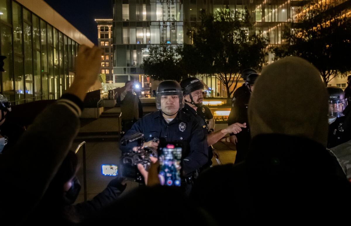 A police officer in a riot helmet stares at a crowd of protesters, with some of them holding phones and cameras to record
