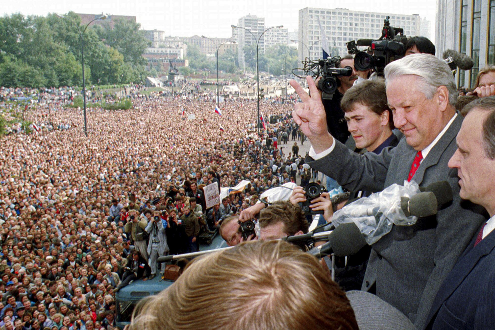 A man, second from right, in a gray suit and red tie makes a V sign as he looks to the masses gathered below him 