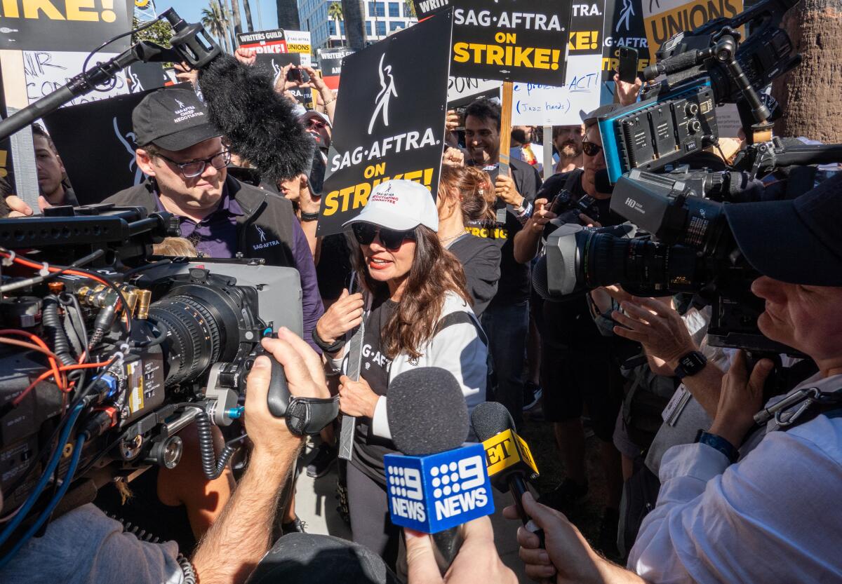 Fran Drescher wears a baseball cap and sunglasses while carrying a picket sign and speaking with reporters at a protest.