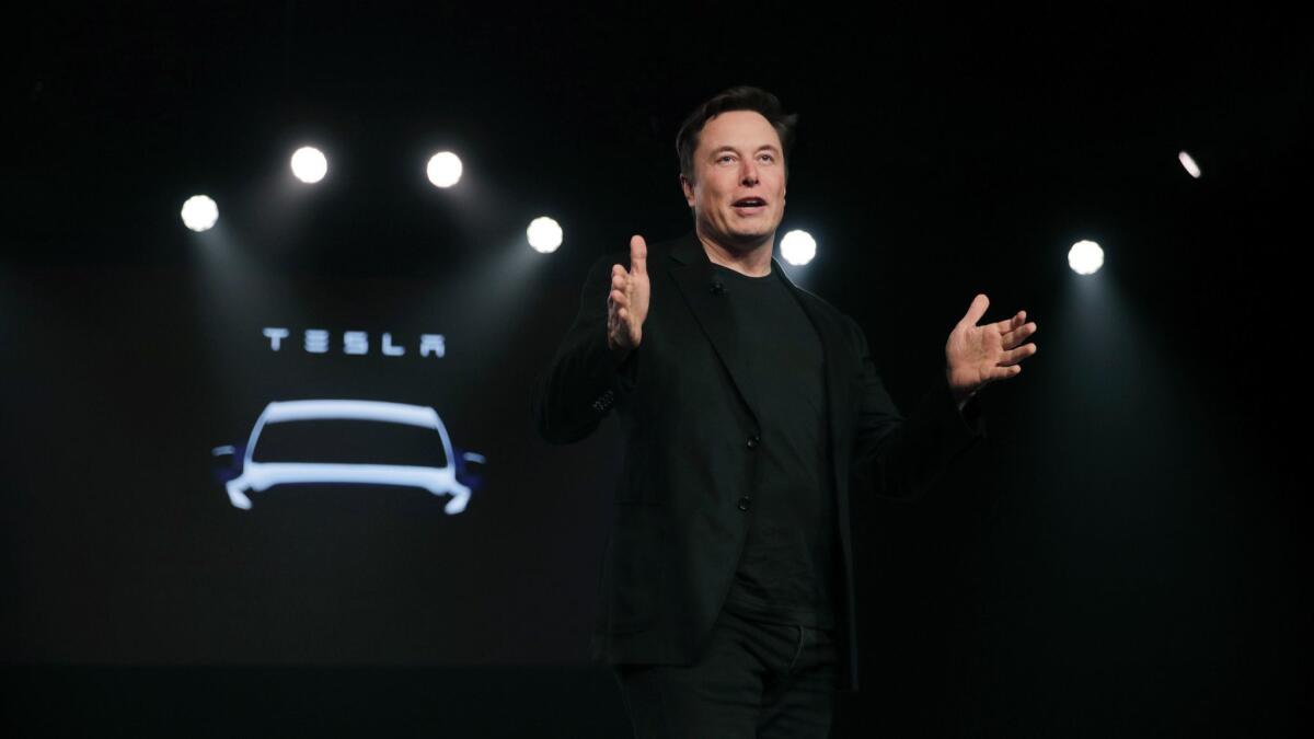Elon Musk stands in front of the Tesla logo, speaking and gesturing with both hands.