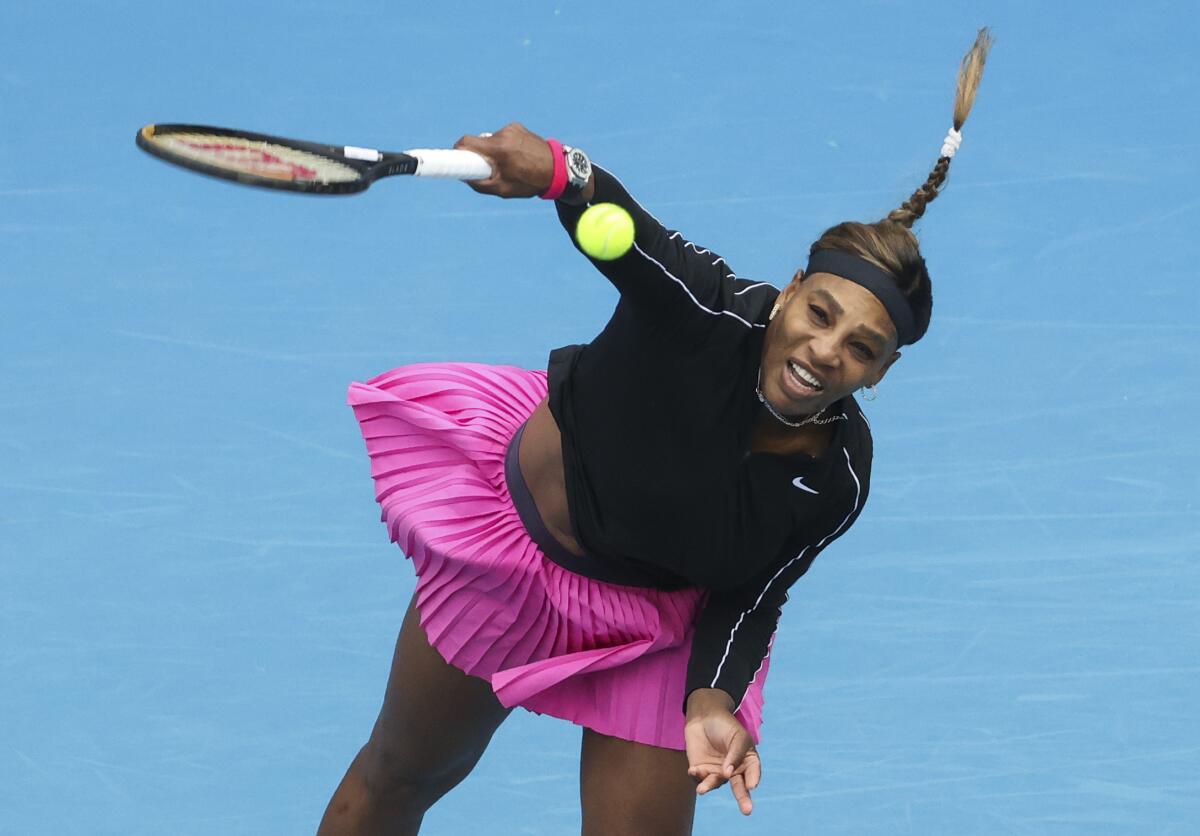 Serena Williams serves the ball while wearing a pink skirt and black top.