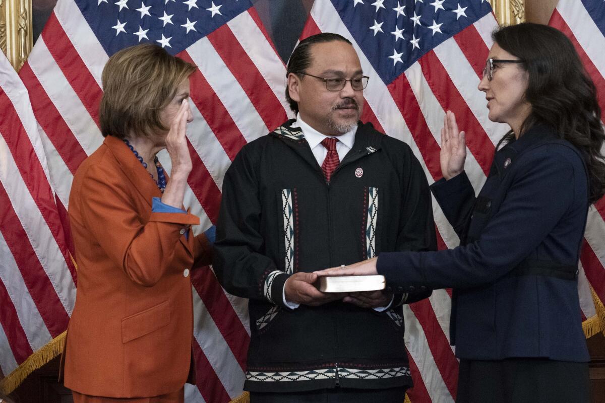 Two women face each other with their right hands raised and a man standing between them on a stage filled with flags