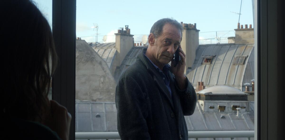 A man stands talking on a cellphone and looking in through a window, with rooftops behind him.