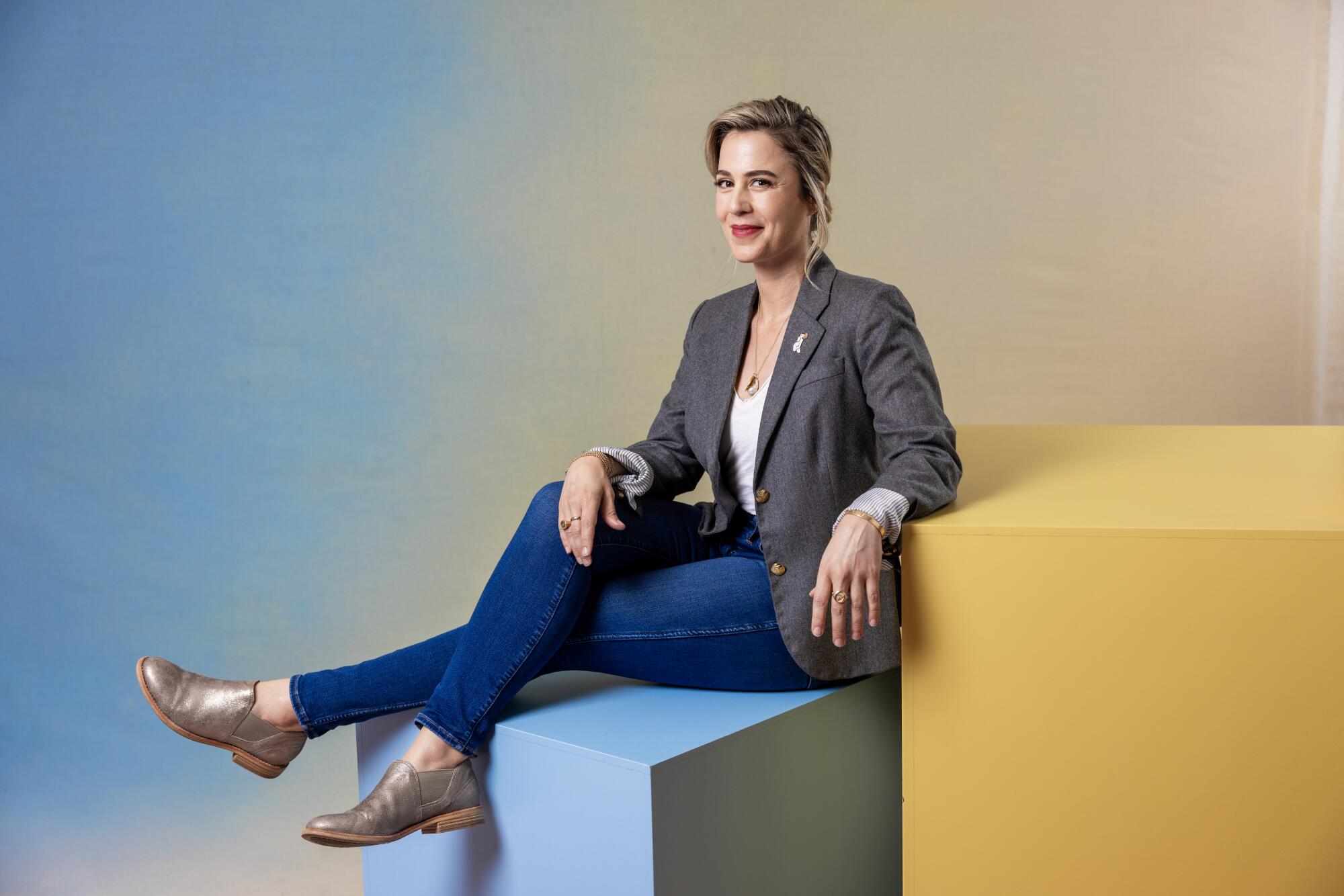 Christy Hall wears a blouse, jacket and jeans while sitting on one cube and resting an arm on another.
