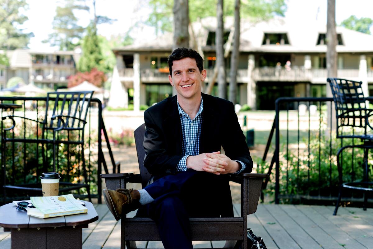 A smiling man sits in an Adirondack chair next to a table with a book and a cup of coffee.