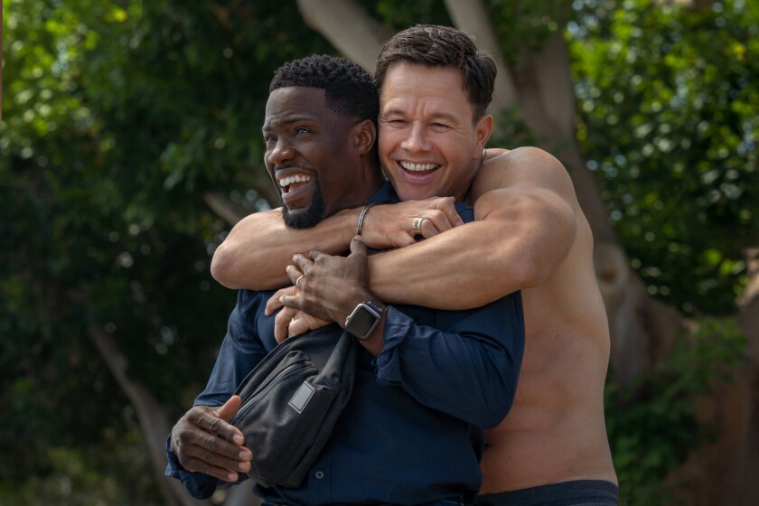 A shirtless man hugs another man from behind.