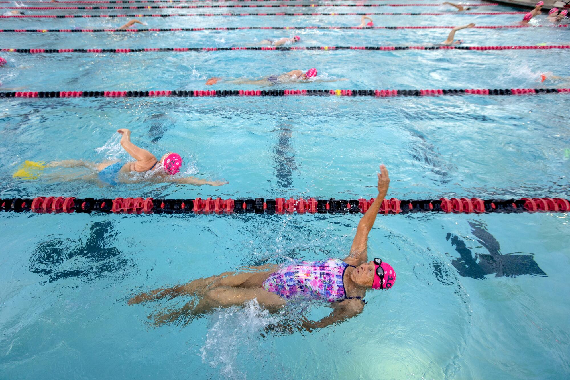 A woman in a pink and blue bathing suit does the backstroke in a pool with other swimmers.