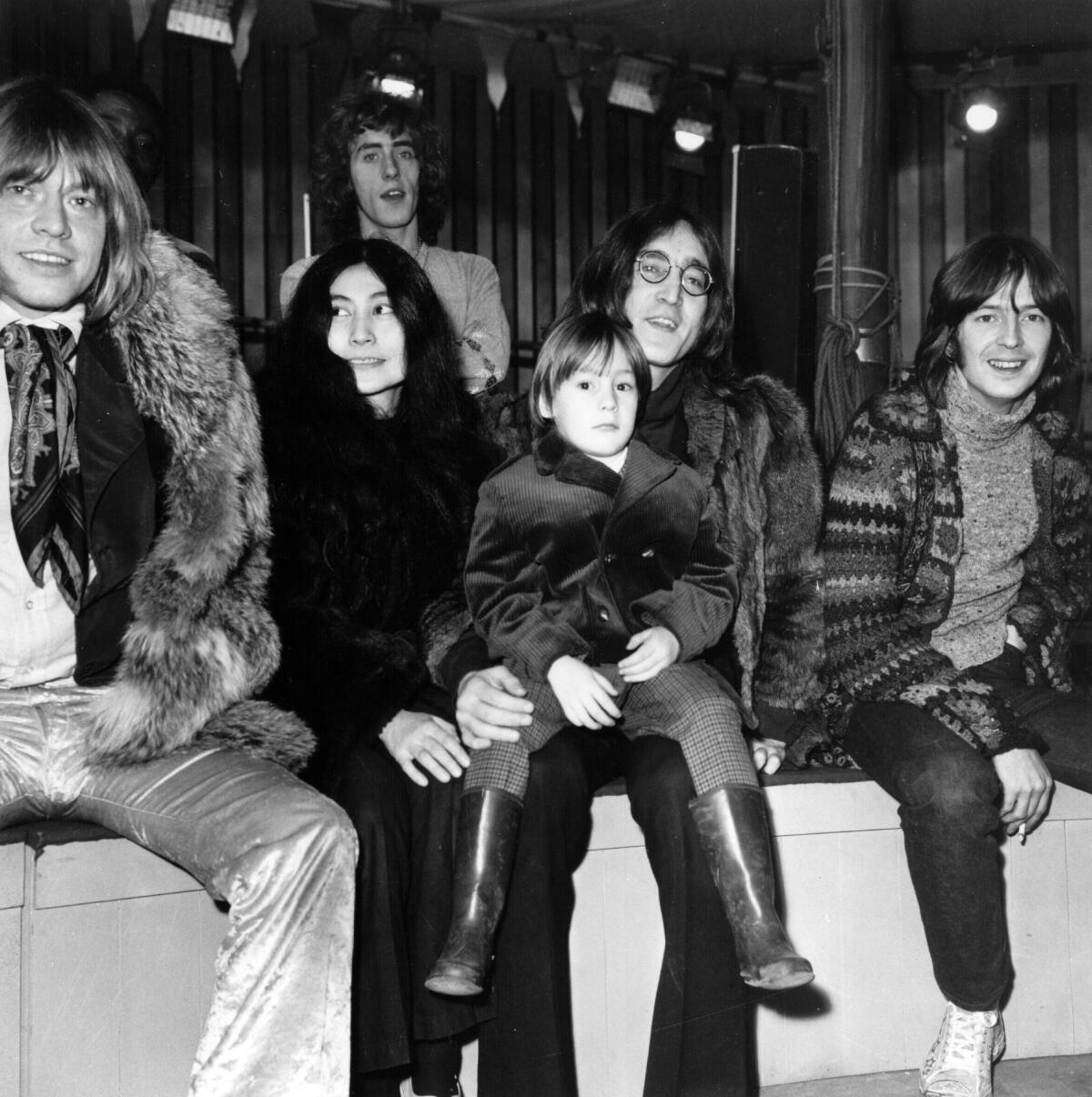 John Lennon, with Yoko Ono and other musicians, sits on a stage with a young Julian Lennon on his lap.