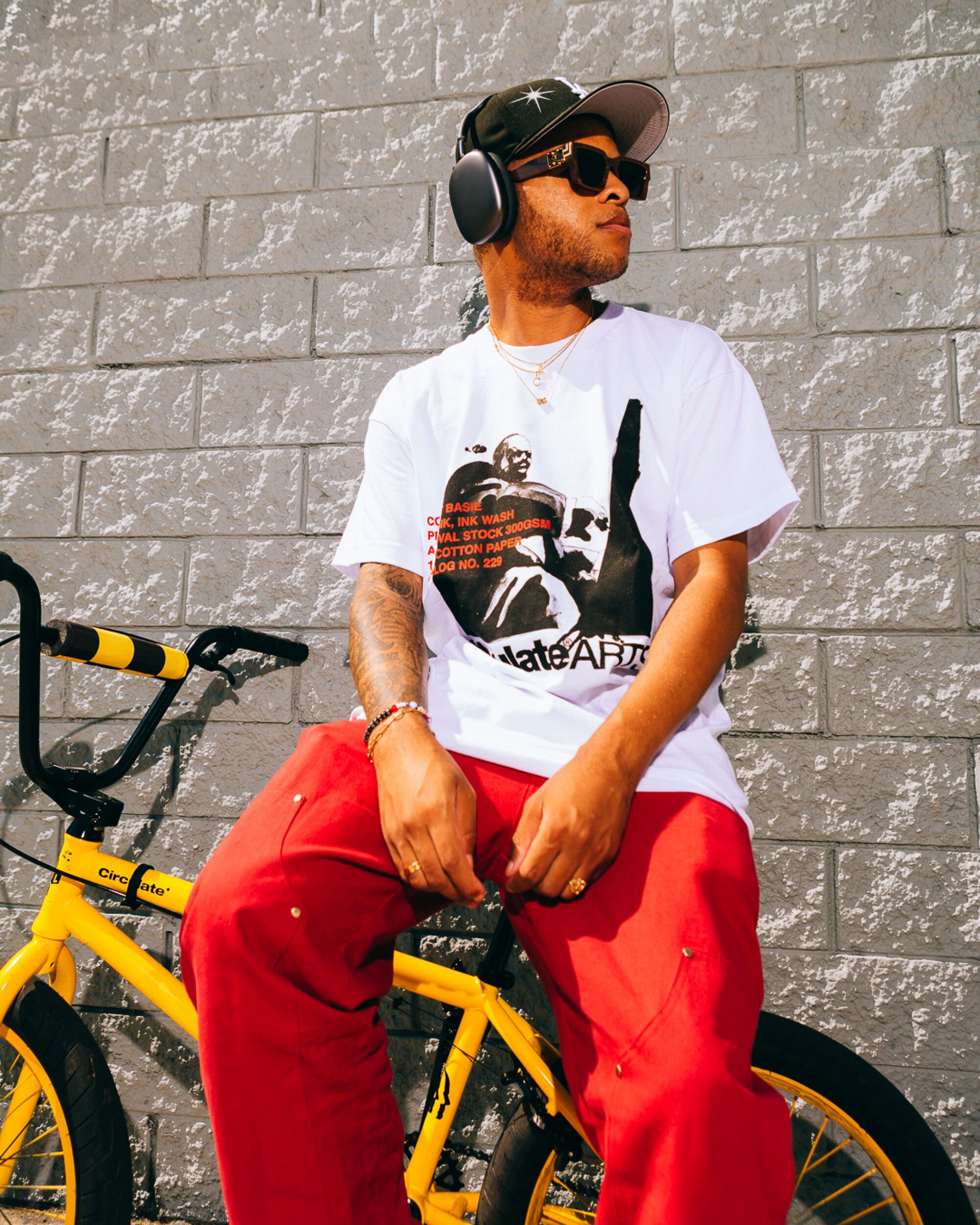 A person in a baseball cap, over-ear headphones, a T-shirt and orange pants leans on a bike against a wall.
