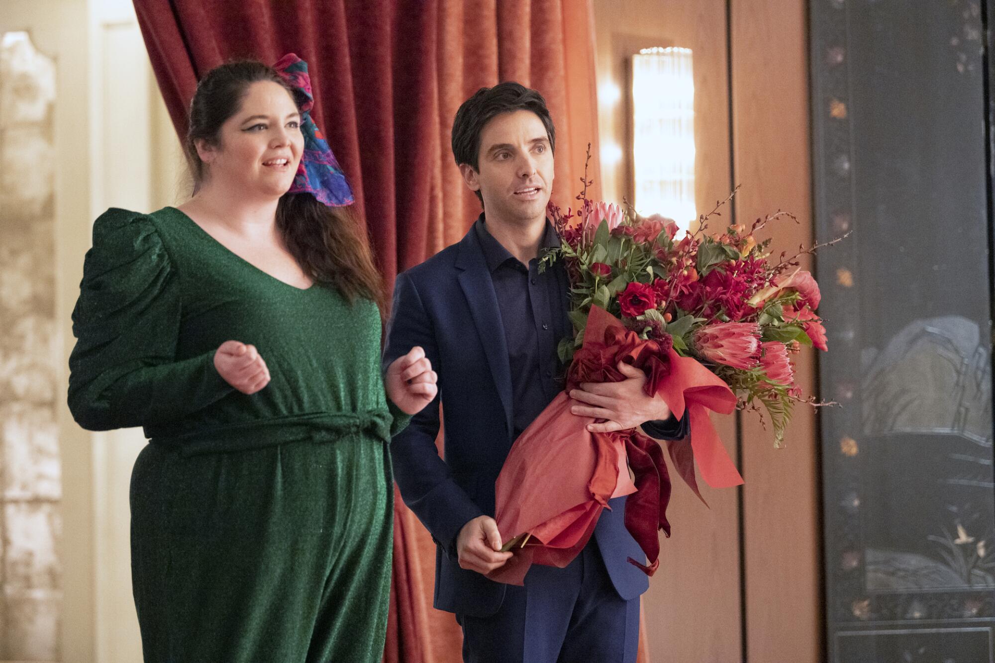 A man carrying a large bouquet of flowers stands with a smiling woman in "Hacks."