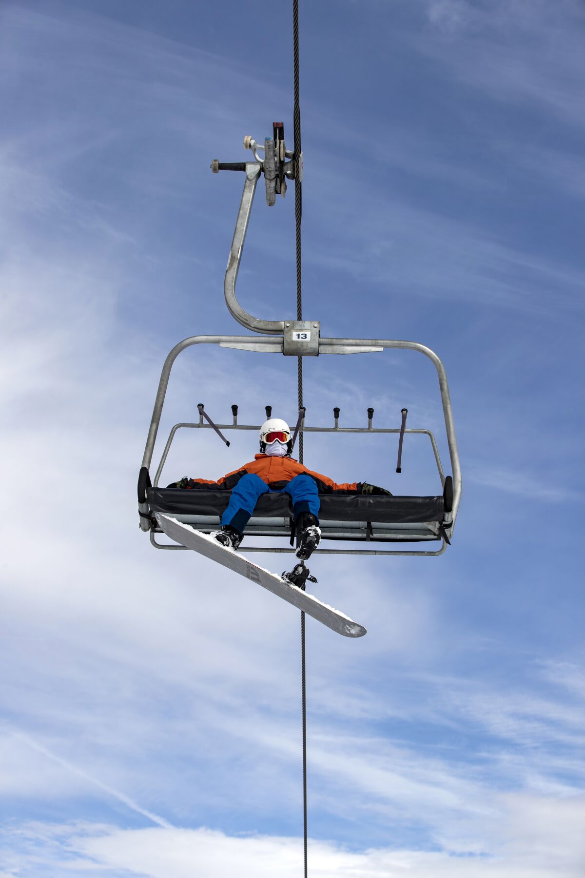 A solo snowboarder, seen from below, rides a chair lift wearing a face covering, snowboard dangling from one foot.