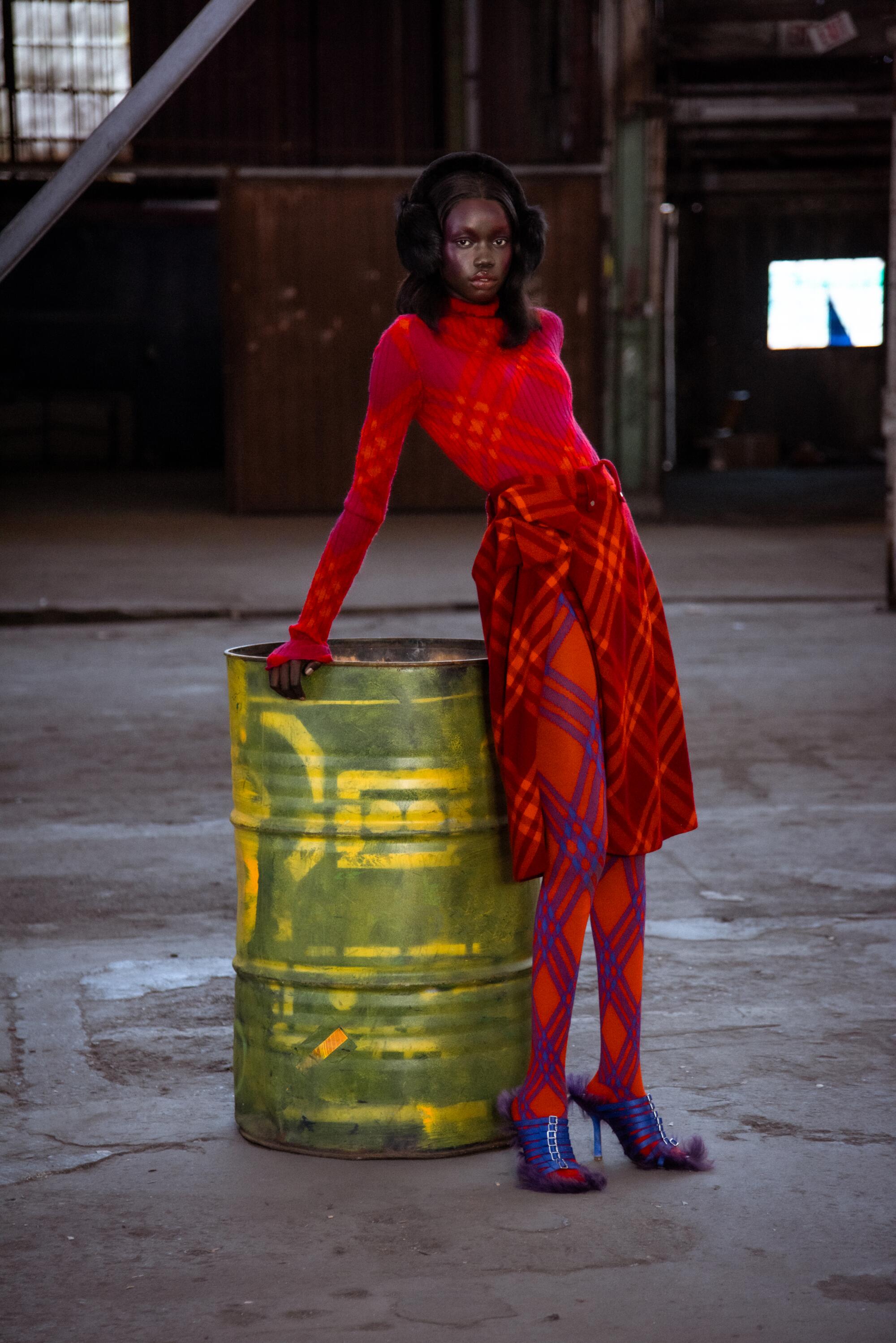 A model in red-orange clothes leans on a bright green trash barrel.