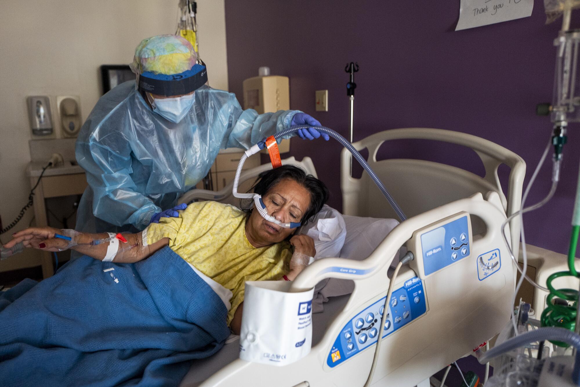 A healthcare worker in personal protective equipment helps a COVID-19 patient with equipment in bed.