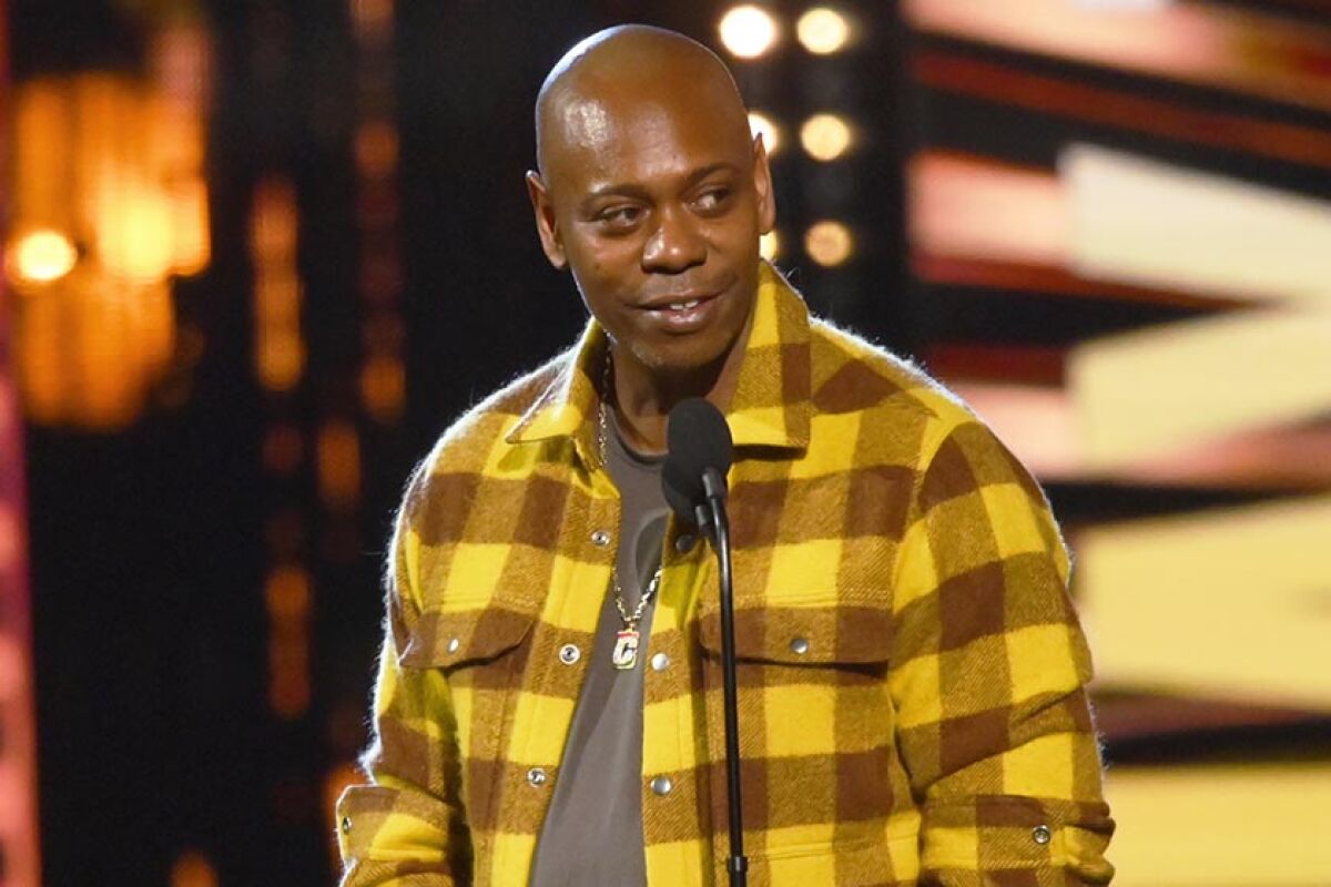 Dave Chappelle stands on stage in front of a microphone.