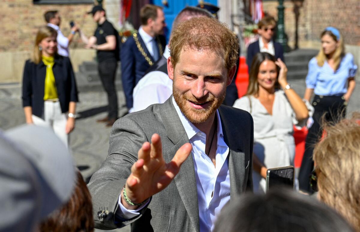 Prince Harry waves to a crowd and smiles in a gray suit jacket and white shirt