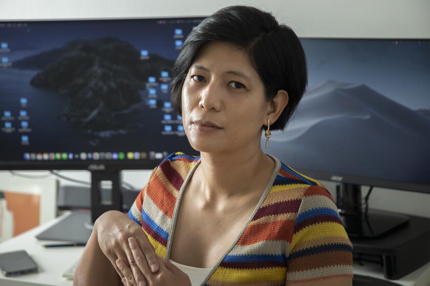 A woman in a striped shirt interlaces her fingers while sitting at a desk with two large computer monitors.