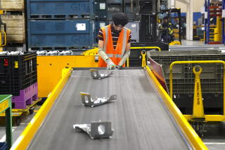 A worker in an orange vest stands at the end of a conveyor belt with metal parts on it