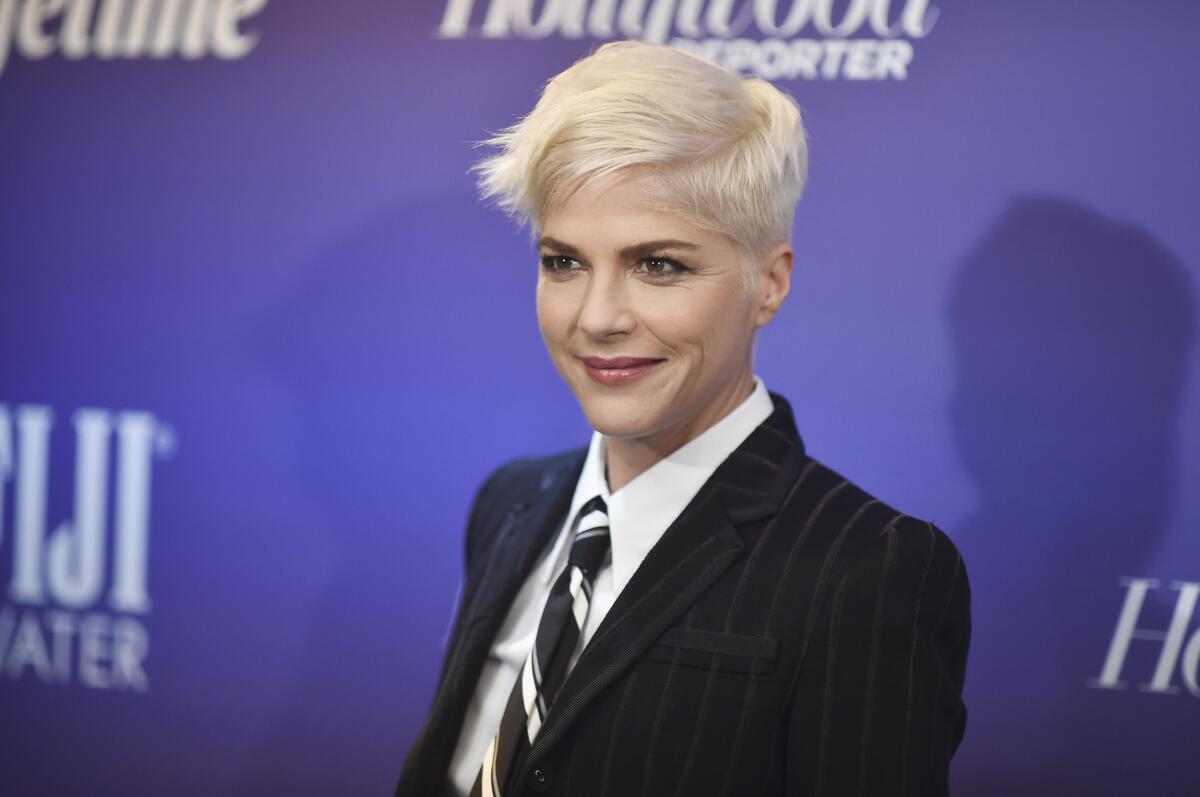 A woman with short blond hair in a dark suit and a striped tie standing in front of a blue backdrop