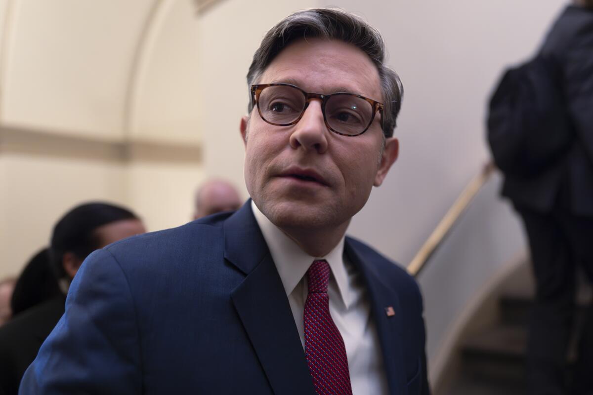 House Speaker Mike Johnson, a man with glasses in a blue suit, turns his head as he walks in a hallway  