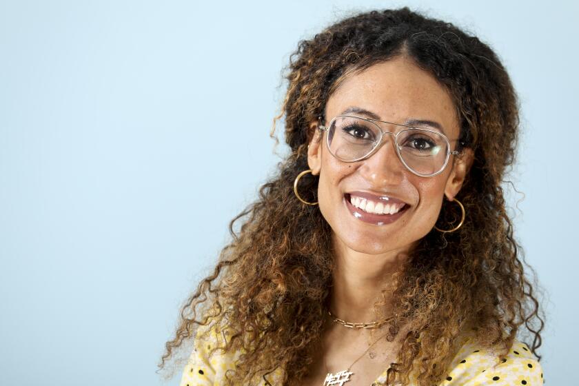 A woman with curly, brown hair and glasses smiling in a yellow polka dot shirt