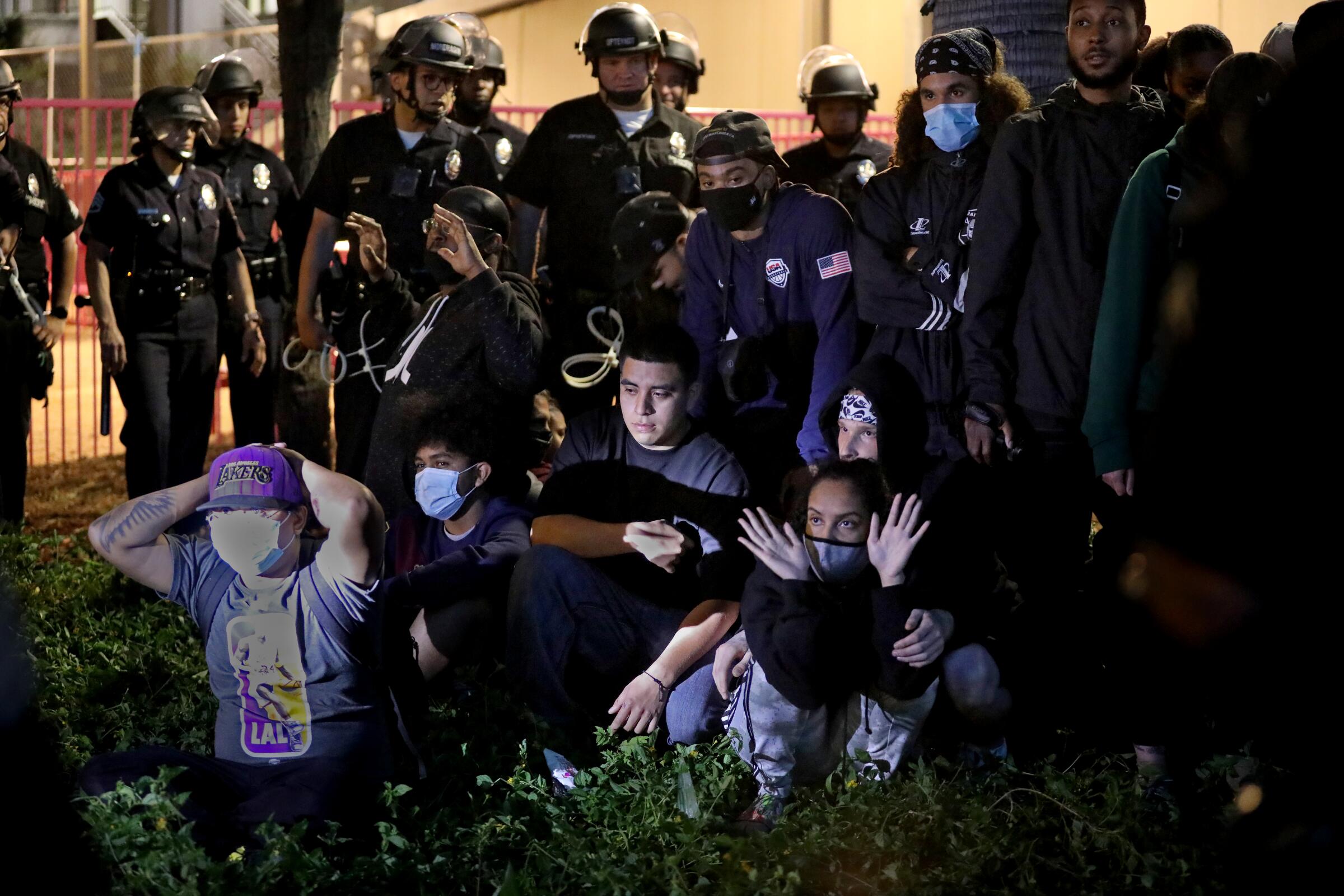 Police in helmets stand behind a group of arrestees who are seated on the ground