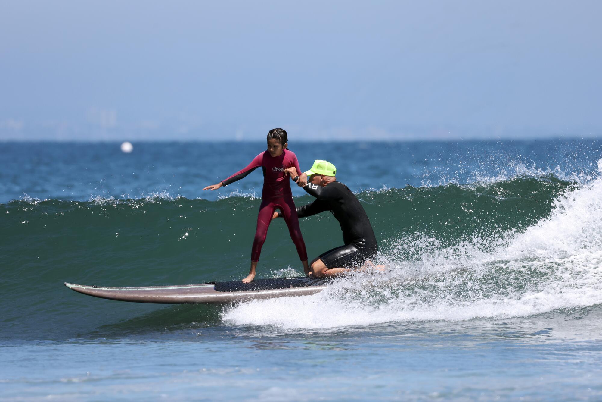 An adult kneels, steadying a young girl, as they ride on a surfboard.