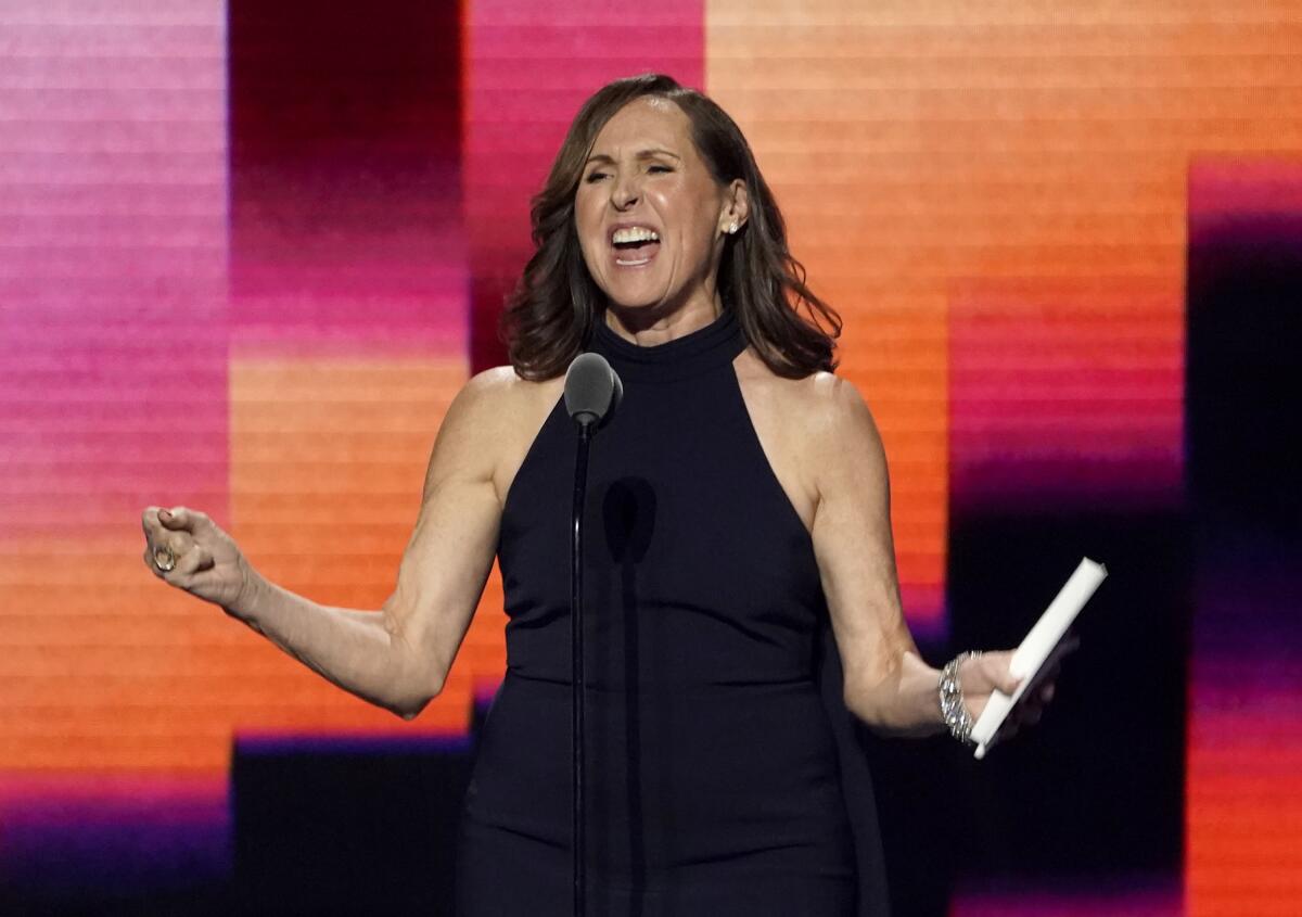 Molly Shannon at a microphone in a black dress.