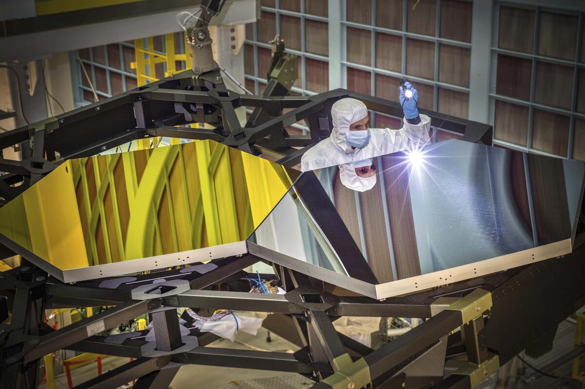 A person in a white suit examines two test mirror segments on a prototype of the James Webb Space Telescope. 