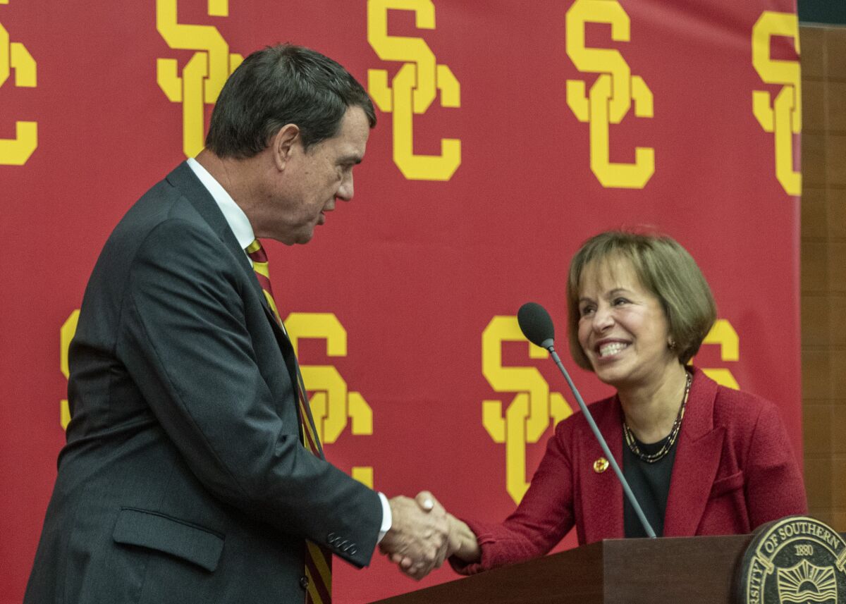 USC athletic director Mike Bohn shakes hands with USC President Carol Folt while standing near a podium.