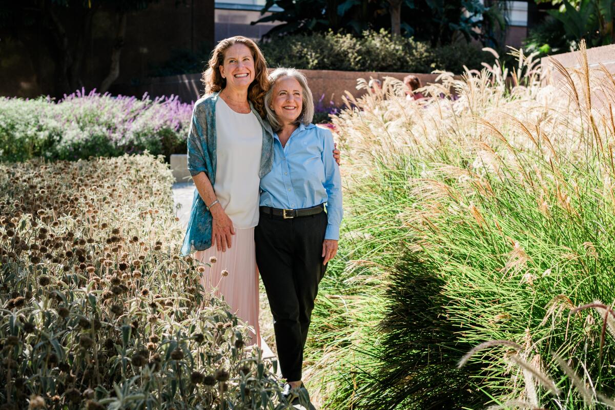 Two women standing arm in arm on a path between ornamental grasses and other plantings